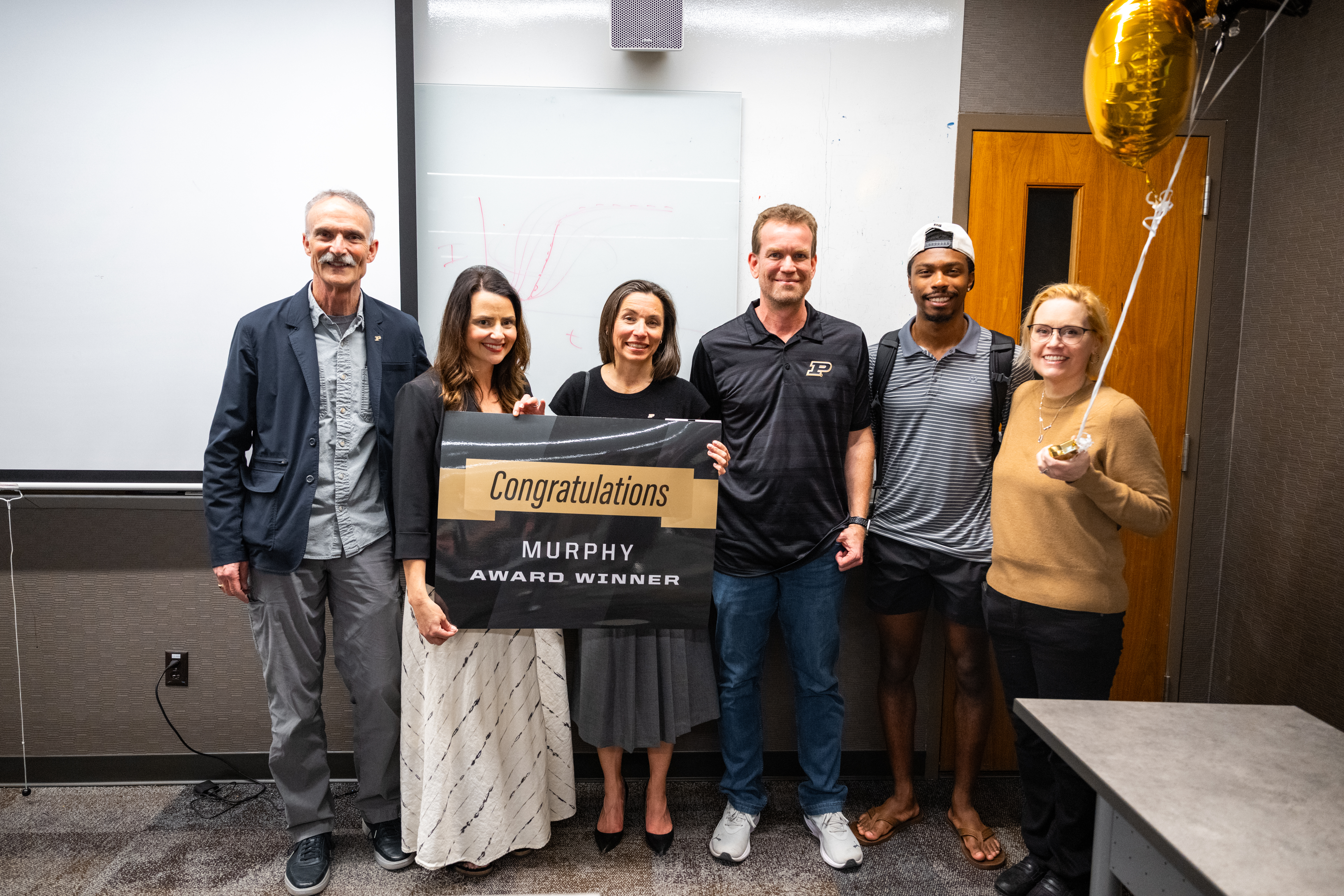 Six people pose together holding a "Congratulations Murphy Award Winner" sign with gold balloons nearby.