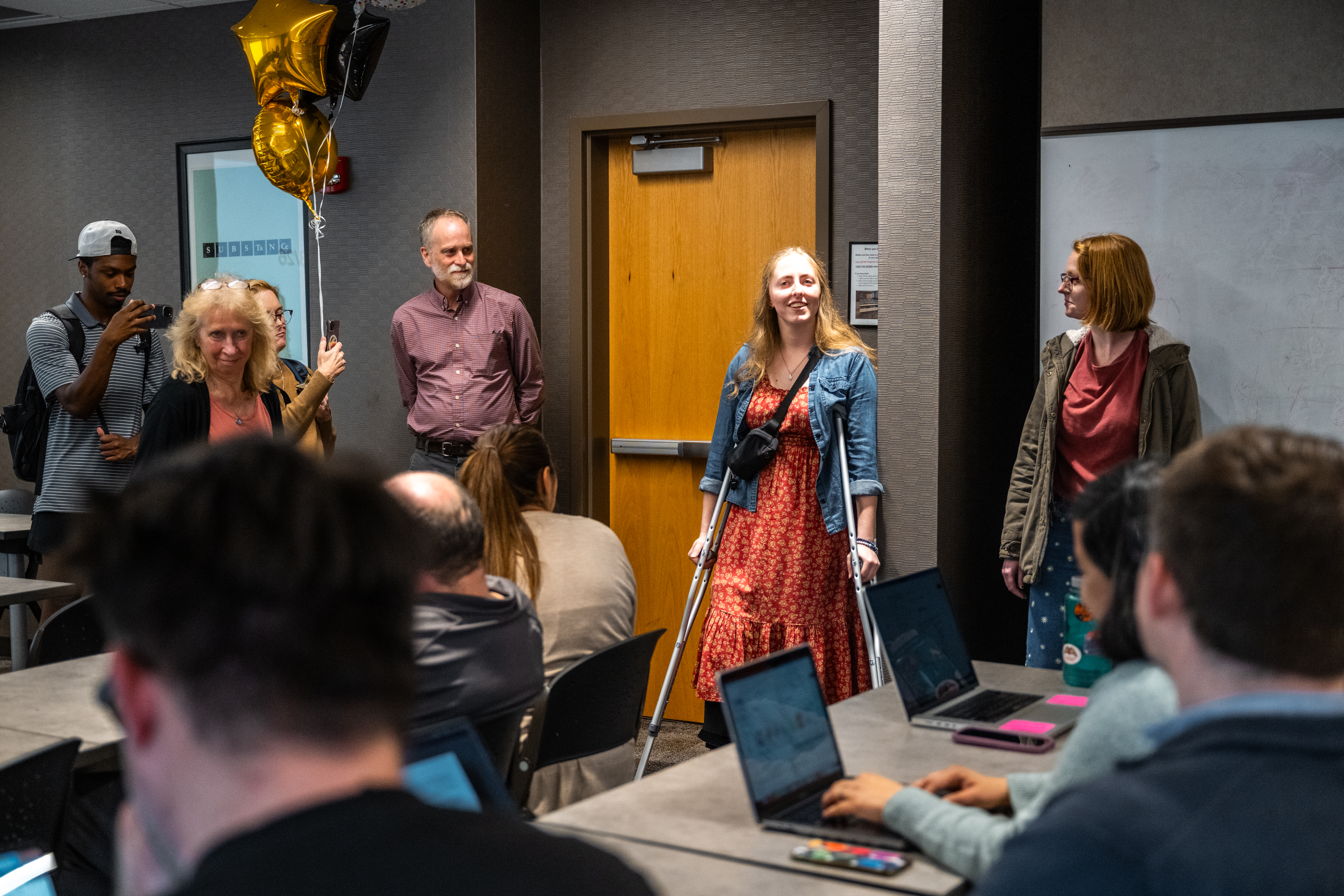 Student Olivia Raab in a red floral dress, on crutches, addresses chemistry faculty at the Murphy Award reveal with Rienstra-Kiracofe.